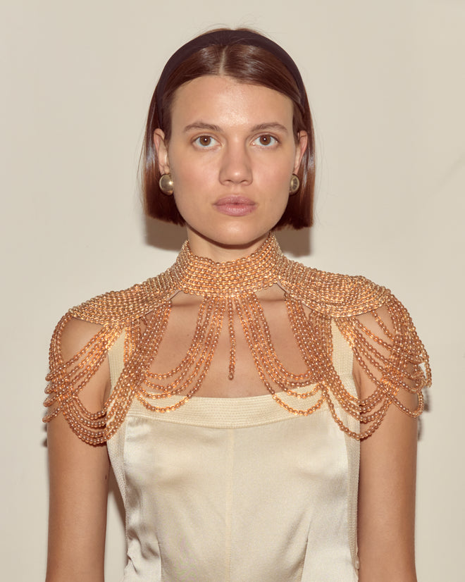 Woman in cream dress with an Epaulette Collar, draped shoulder jewelry, and pearl earrings stands against a plain background. Subtle 1960s-inspired accessories complete her timeless look.