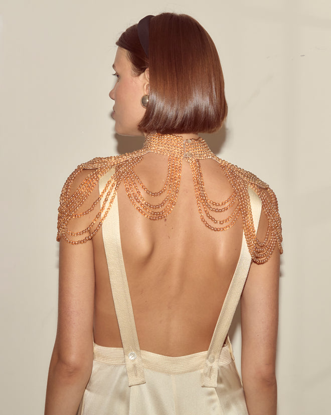A woman with short brown hair, facing away, wears the Epaulette Collar beaded shoulder chain—reminiscent of 1960s accessories—with a backless cream outfit.