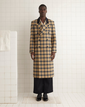 A person wears the Equestrian Mitsy Coat, a double-breasted plaid wool coat with black lapels, crafted from classic British mill fabric, standing stylishly against a tiled background.