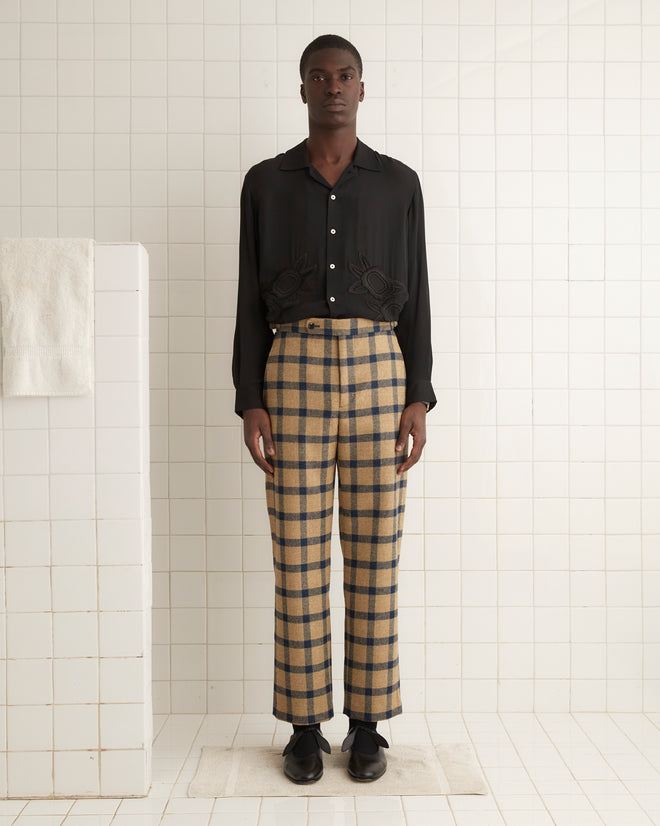 A man in a plaid suit sits on a bench, his Equestrian Trousers with side-buckle waist adjusters providing a striking contrast against the dark blue wall and vivid orange floor.