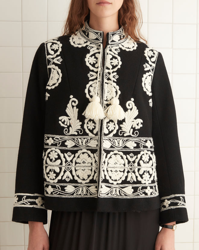 A woman stands in a tiled room wearing a black Estate Jacket, a black fringe skirt, and black shoes with ankle straps.