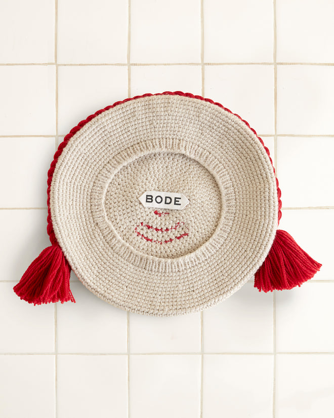 A Face Beret features a round woven design with googly eyes, a red yarn smile, and braided red yarn hair on a tiled background, charmingly reminiscent of a playful mat.