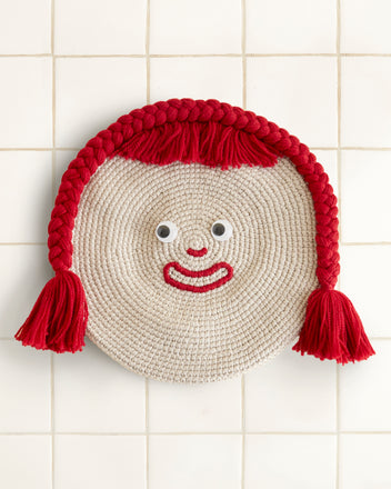 A Face Beret features a round woven design with googly eyes, a red yarn smile, and braided red yarn hair on a tiled background, charmingly reminiscent of a playful mat.