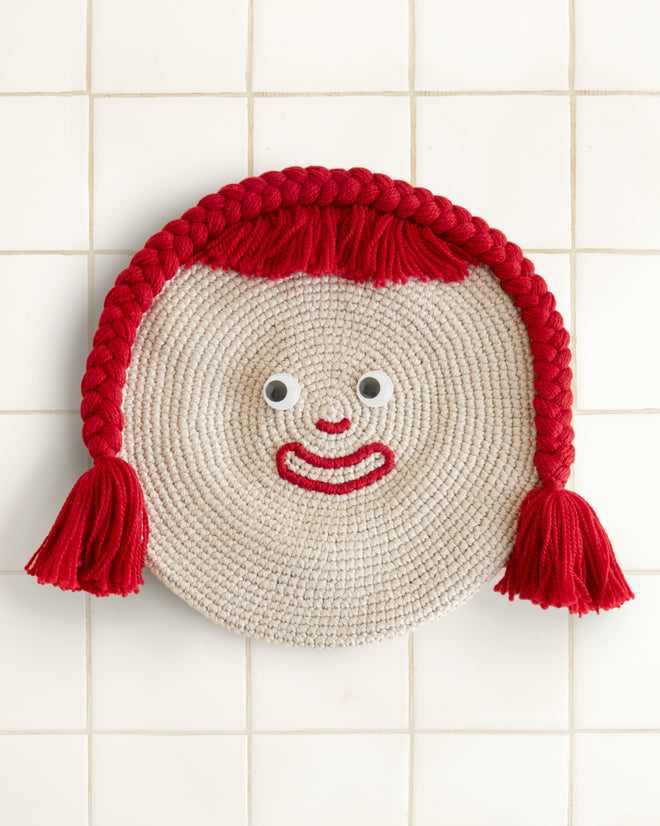 A Face Beret features a round woven design with googly eyes, a red yarn smile, and braided red yarn hair on a tiled background, charmingly reminiscent of a playful mat.