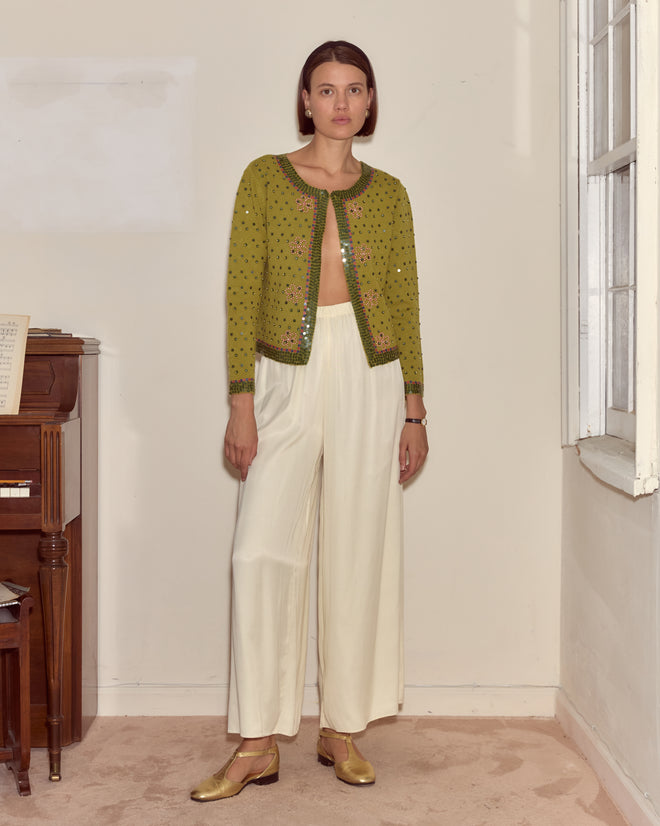 A woman stands indoors wearing the Fairyfloss Cardigan, a green sequin piece with beaded floral appliqué, paired with white wide-leg pants and gold shoes.
