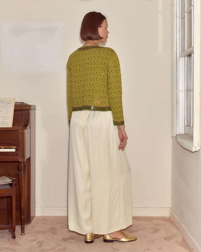 A woman wears the Fairyfloss Cardigan, a green wool-cashmere knit, paired with white wide-leg pants, standing indoors by a piano and window.