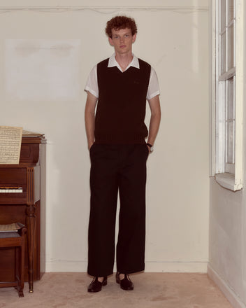 A man wearing the Falls Vest - Dark Brown with dark pants and a white shirt stands indoors near a piano and window.
