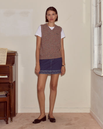 A woman stands indoors by a piano, wearing the Falls Vest - Multi from the latest women's knitwear collection, styled with a white shirt, denim skirt, and loafers.
