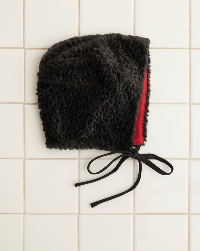 A person in a green knit outfit, enhanced by chic accessories like the Faux Fur Tie Cap in black, poses against a red curtain backdrop.