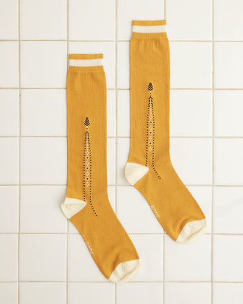 The Geo Diamond Sock, with its vibrant yellow hue and a modern interpretation of 1930s-1940s style, boasts a vertical black dotted geometric pattern along with contrasting white toe and heel. Crafted from 100% cotton, these knee-high socks are elegantly displayed on a pristine white tiled floor.