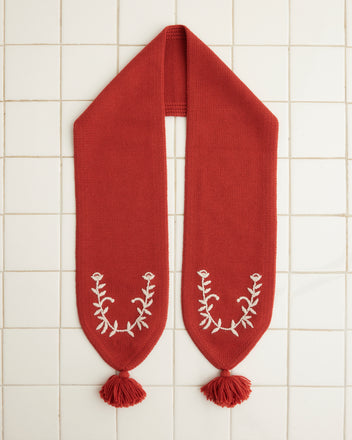 The Floret Scarf - Red, crafted from merino wool and featuring white leaf embroidery with tassels, is elegantly displayed on a tiled surface.