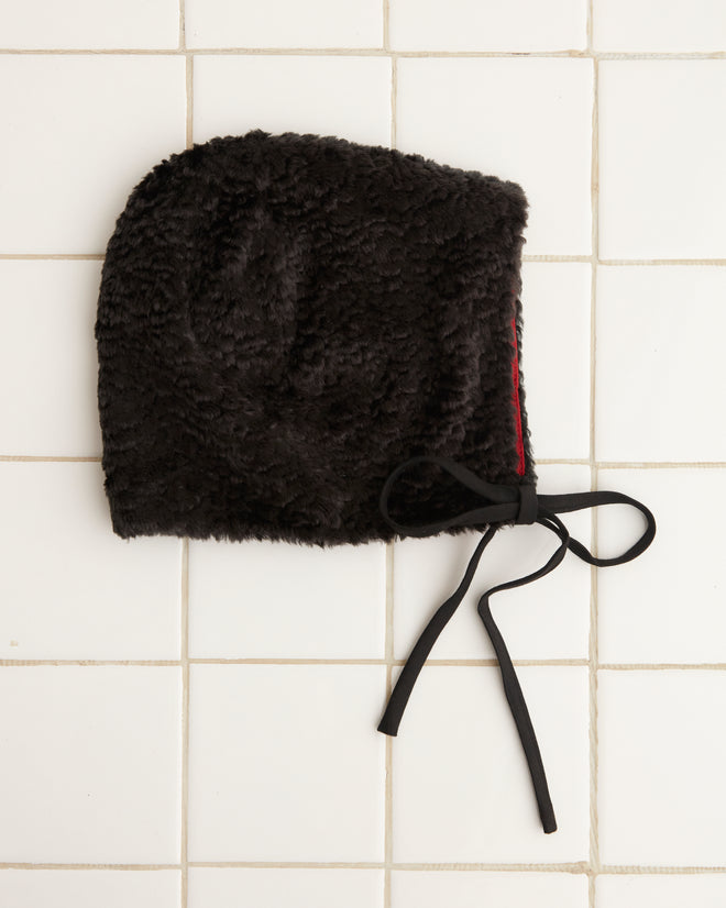 Close-up of the Faux Fur Tie Cap - Black with a red lining, made from polyester, placed on a light tiled surface.