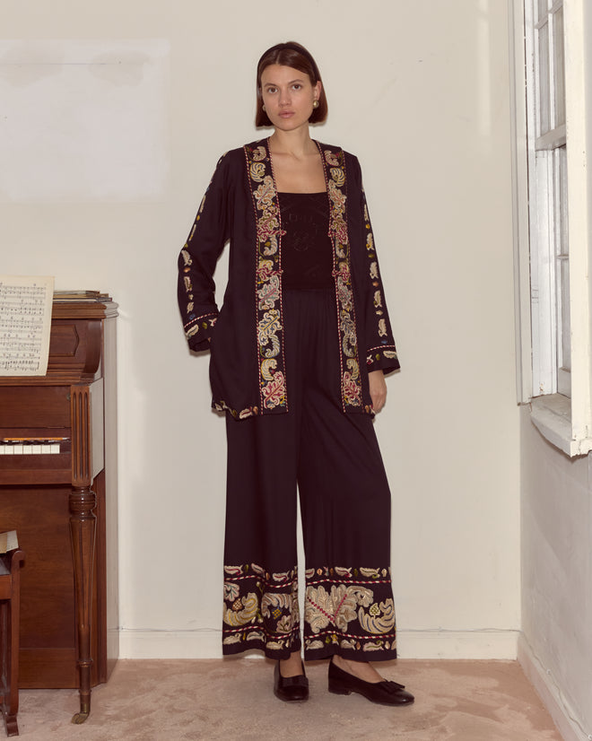 Woman in a Feather Runner Jacket, inspired by the Georgian era with black hand embroidery, stands indoors next to a piano and looks at the camera.