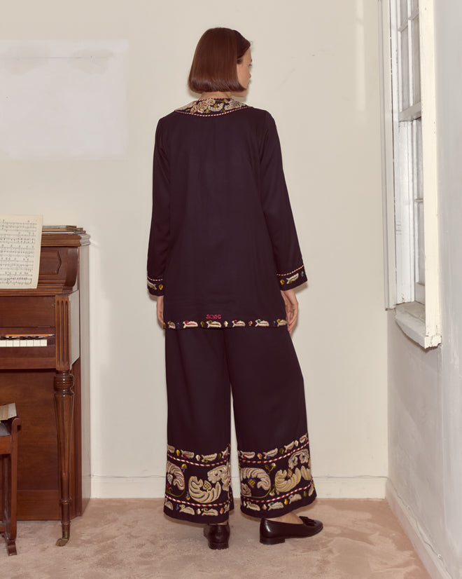A woman wearing the Feather Runner Jacket in black, featuring hand embroidery inspired by the Georgian era, stands by a piano in a softly lit room, facing away.