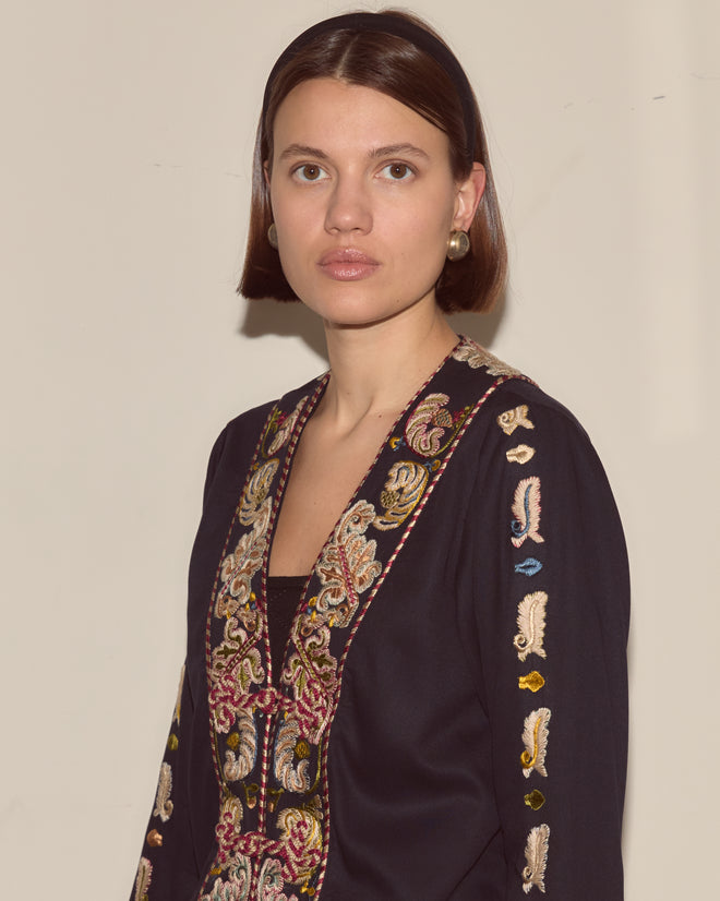 Woman with straight brown hair facing forward, wearing the Feather Runner Jacket—a dark, hand-embroidered Georgian era reproduction—and pearl earrings.