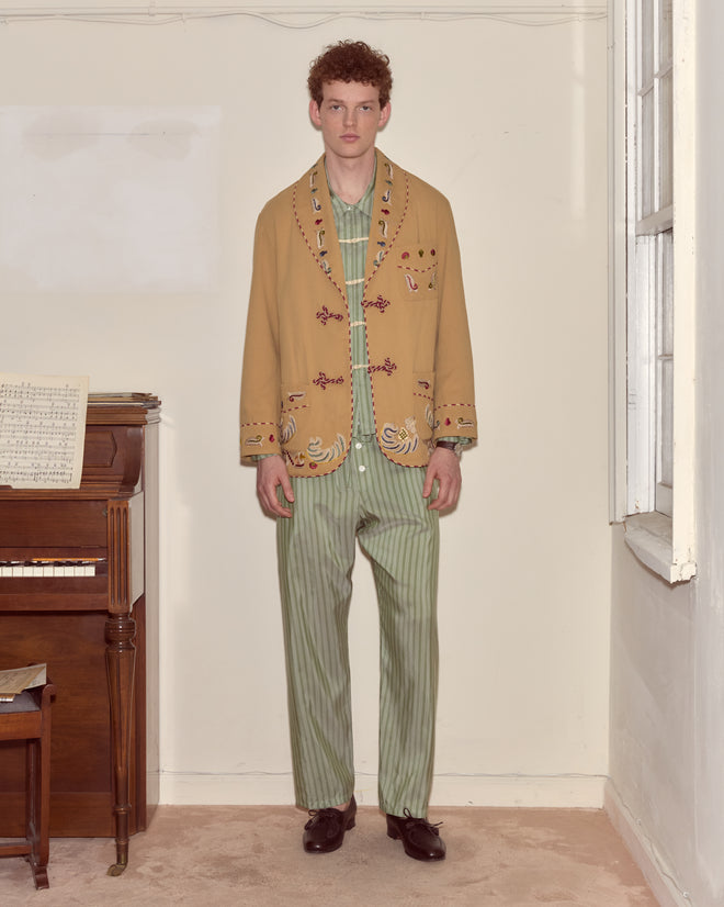 A man in striped green pajamas and a Feather Runner Smoking Jacket stands by a piano in a sparsely furnished room.