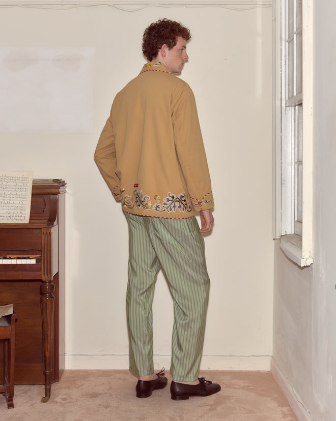 A man in the Feather Runner Smoking Jacket with Georgian-era embroidery and striped green pants stands by a piano, facing away in a bright room.