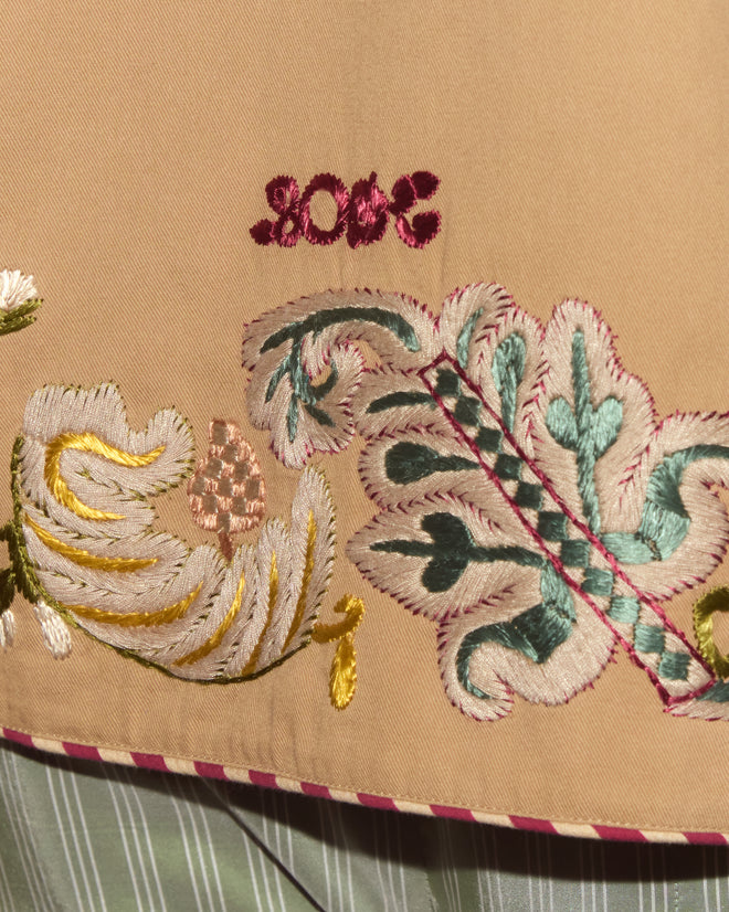 Close-up of hand embroidery on the Feather Runner Smoking Jacket, showcasing pastel floral patterns on beige fabric in a Georgian style, set against a striped green background.
