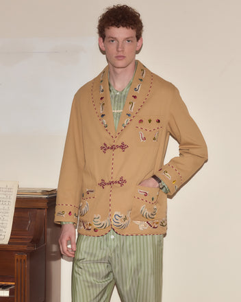 A man in a Feather Runner Smoking Jacket with Georgian-era hand embroidery and green striped pants stands by a piano with sheet music.