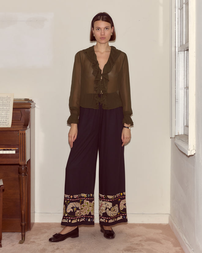 Woman in a sheer olive blouse and Feather Runner Pants - Multi, featuring wide legs and colorful hand embroidery at the hem, standing by a piano.