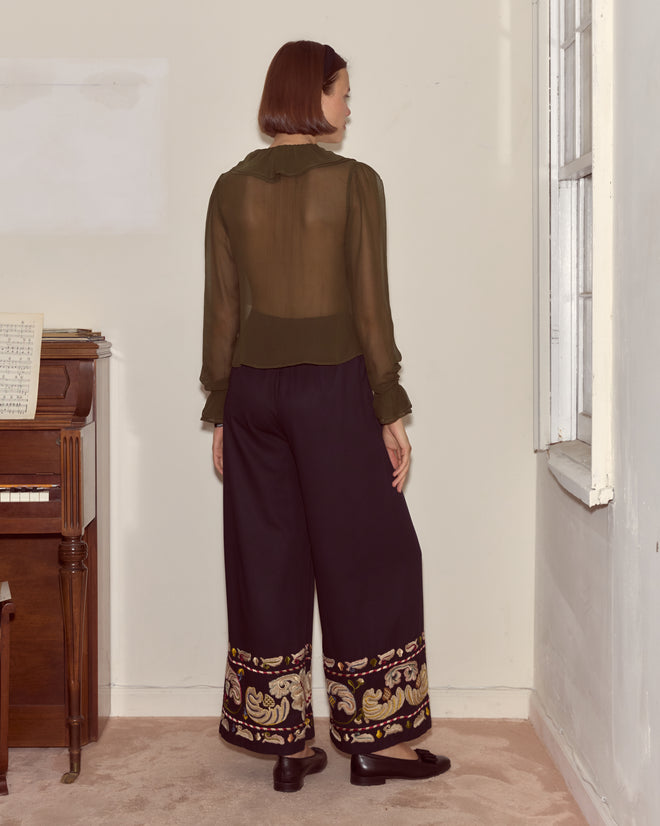 A woman in sheer olive, hand-embroidered blouse and Feather Runner Pants – Multi stands facing a wall beside a piano.