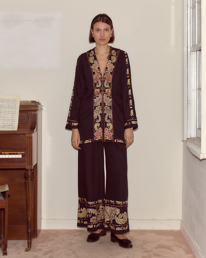 A woman wearing the Feather Runner Jacket—black, gold, and red with hand-embroidered Georgian era details—stands indoors beside a piano and window.