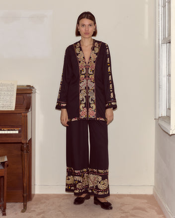 A woman wearing the Feather Runner Jacket—black, gold, and red with hand-embroidered Georgian era details—stands indoors beside a piano and window.