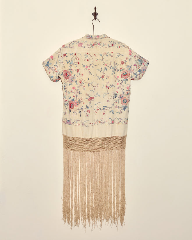 The Festival Blossom Tunic - S is a unique floral short-sleeve shirt with long beige fringe, crafted from an antique piano shawl and displayed on a wall hook.