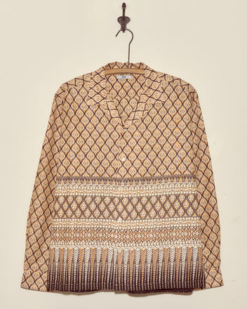 The Fictile Flame Long Sleeve Shirt (L/XL), featuring a unique brown wax-print pattern, hangs on a hanger against a plain wall.