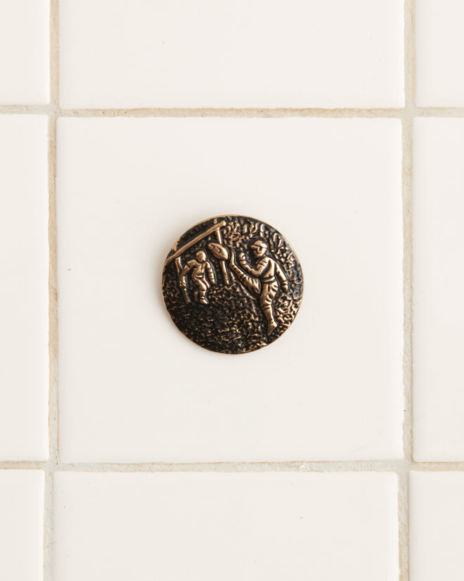 A Field Goal Brooch, a round, bronze-colored coin featuring a relief of two figures in an outdoor scene reminiscent of an early 20th century football pin, is placed on a white tiled surface.