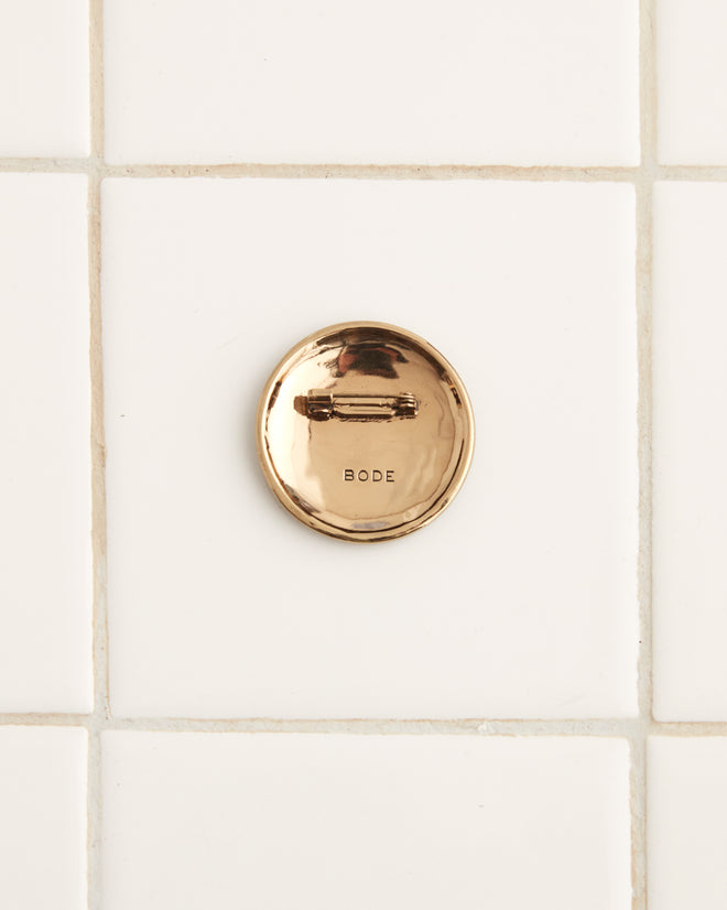 A Field Goal Brooch, a round, bronze-colored coin featuring a relief of two figures in an outdoor scene reminiscent of an early 20th century football pin, is placed on a white tiled surface.