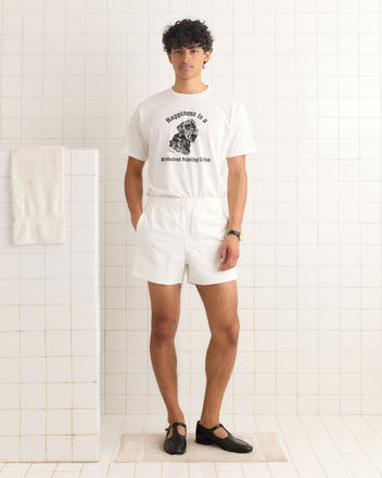 A man dressed in all-white, including Field Shorts - White, stands on a bath mat in a tiled bathroom with a towel hanging nearby.