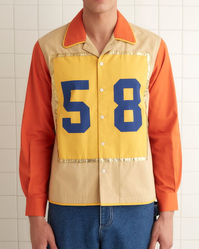 A person wearing the Fifty-Eight Shirt, featuring yellow and orange colors with metallic gold trim, paired with blue jeans and black shoes, stands against a tiled wall.