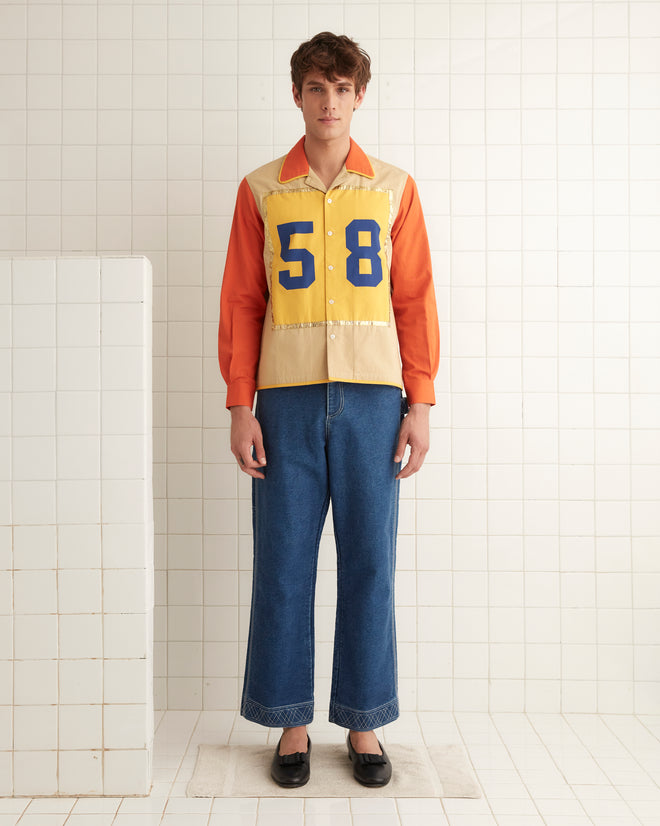 A person wearing the Fifty-Eight Shirt, featuring yellow and orange colors with metallic gold trim, paired with blue jeans and black shoes, stands against a tiled wall.