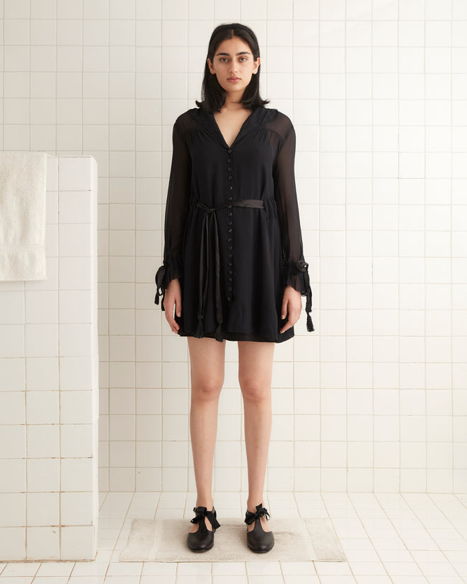 In a white-tiled room, an individual is wearing the Finch Dress in black, which has long sleeves tied at the wrists and an unfitted silhouette, paired with black shoes.