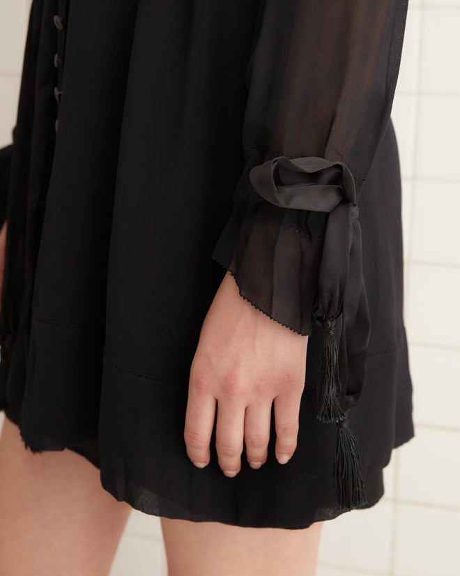 In a white-tiled room, an individual is wearing the Finch Dress in black, which has long sleeves tied at the wrists and an unfitted silhouette, paired with black shoes.