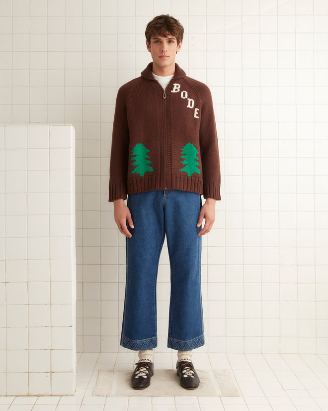 The individual sports a vintage Fir Shawl Collar Cardigan by BODE, adorned with green trees and crafted from 100% wool. Complementing the cozy knit are blue jeans and brown shoes, creating a stylish indoor ensemble.