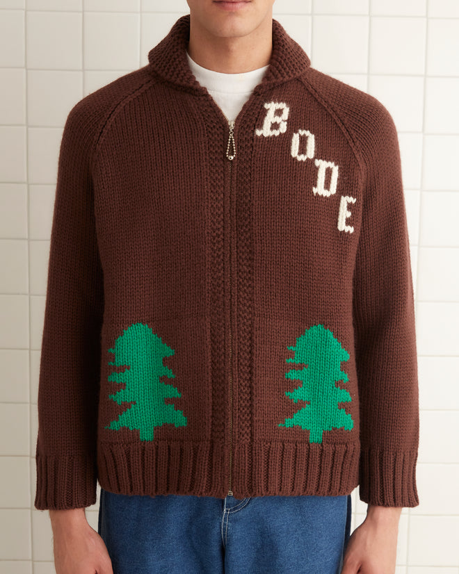 The individual sports a vintage Fir Shawl Collar Cardigan by BODE, adorned with green trees and crafted from 100% wool. Complementing the cozy knit are blue jeans and brown shoes, creating a stylish indoor ensemble.