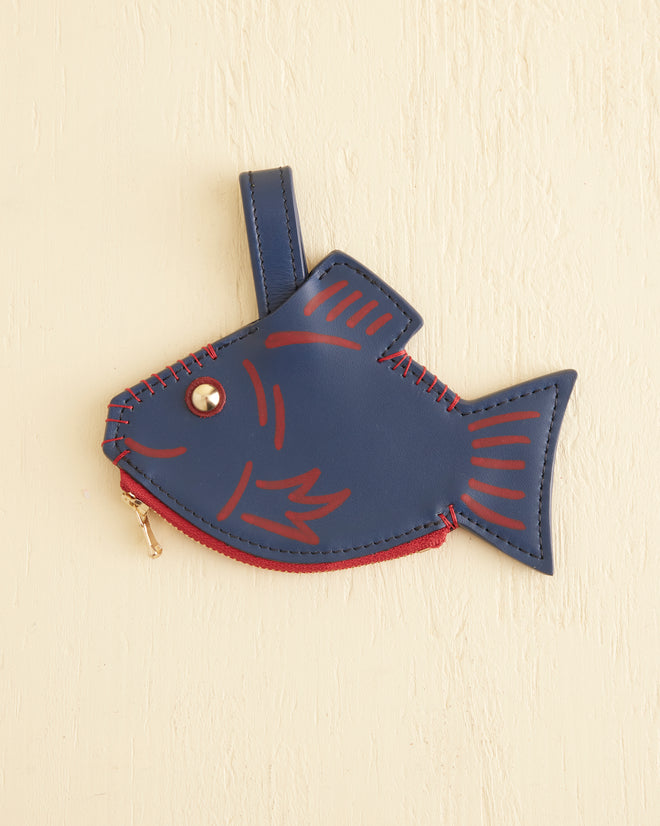 A Fish Coin Purse, featuring a blue and red vintage design with a metal zipper, hangs against a beige textured wall.