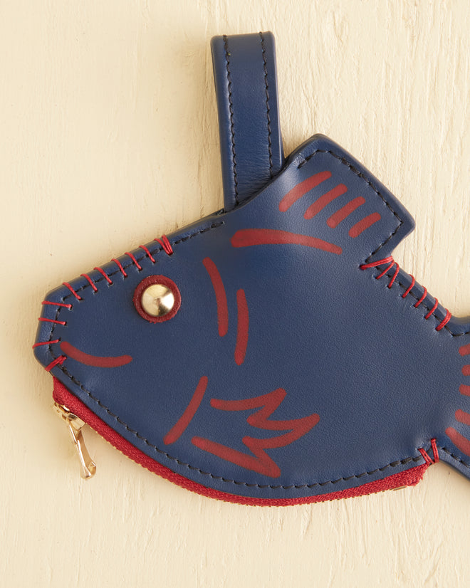 A Fish Coin Purse, featuring a blue and red vintage design with a metal zipper, hangs against a beige textured wall.