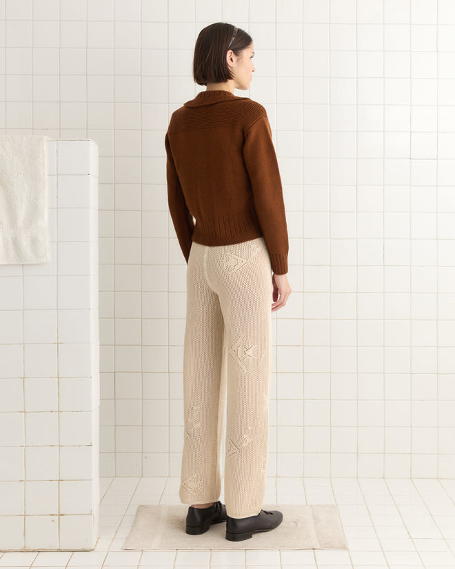 In a tiled room, a person with short hair wears a brown sweater knit lace and cream Angel Fish Joggers, made of 100% cotton, paired with dark shoes.