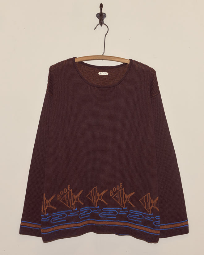 The Fish School Sweater is a brown knit cotton sweater featuring blue and tan fish and wave patterns along the bottom, displayed on a wooden hanger—a cozy nod to angel fish style.
