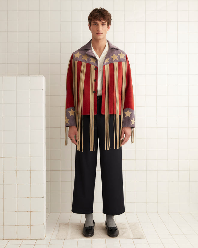 Dressed in the eye-catching Flag Fringe Jacket, featuring stars and long fringes, a person confidently stands in a tiled room. They pair the jacket with a white shirt, black pants, and black shoes.