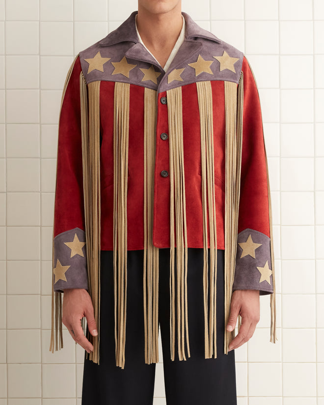 Dressed in the eye-catching Flag Fringe Jacket, featuring stars and long fringes, a person confidently stands in a tiled room. They pair the jacket with a white shirt, black pants, and black shoes.