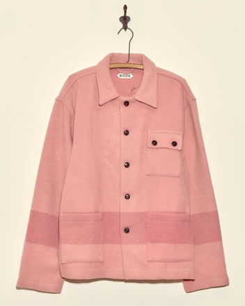 The Flamingo Pink Line Jacket (XL/XXL), a unique midcentury wool piece with black buttons and front pockets, hangs from a simple hook on a beige wall.