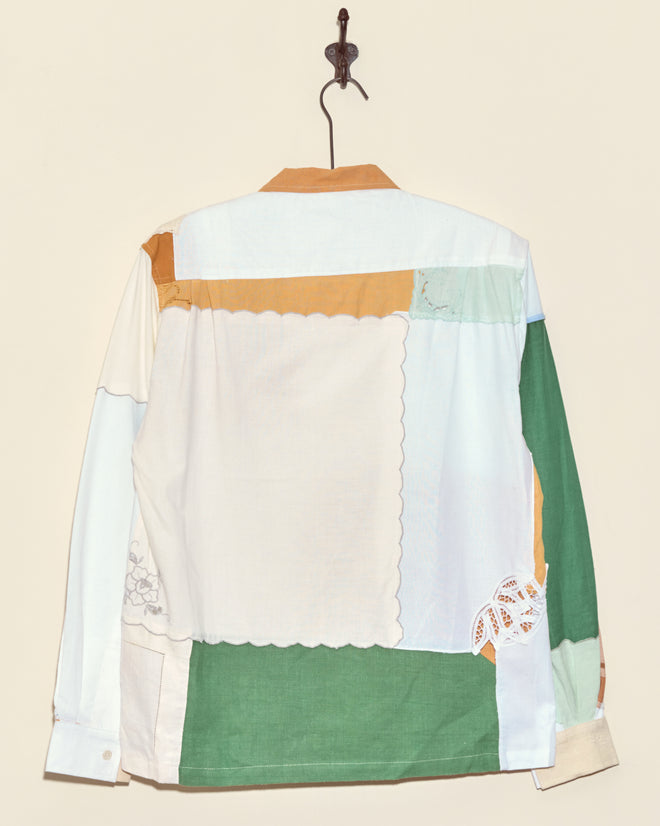 The Flaming Petal Napkin Shirt - S/M, featuring unique patchwork in green, white, blue, and tan fabrics, hangs on a wall hook.