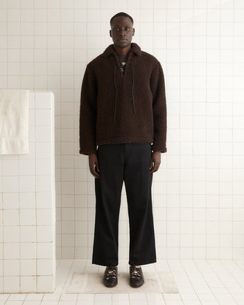 A person in a dark brown, long-sleeve Fleece Tie-Up Pullover with black pants and brown loafers stands against a plain, light-colored wall on a carpet with a floral pattern.