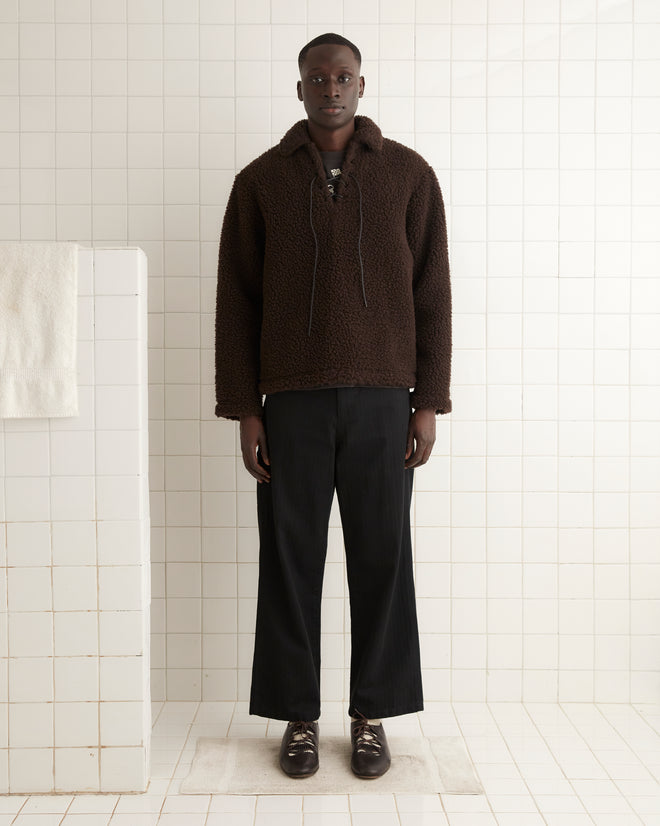 A person in a dark brown, long-sleeve Fleece Tie-Up Pullover with black pants and brown loafers stands against a plain, light-colored wall on a carpet with a floral pattern.