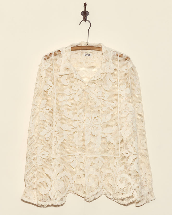 The Fleur Lace Long Sleeve Shirt - L is a cream, sheer crochet button-up with unique floral patterns and filet lace details, displayed hanging against a beige wall.