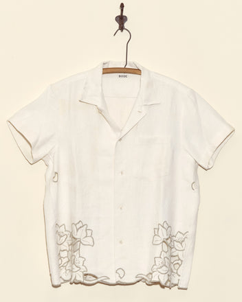 Floating Lily Short Sleeve Shirt - M: White short-sleeve button-up with floral embroidery along the hem, crafted from vintage textile, displayed hanging on a hook.